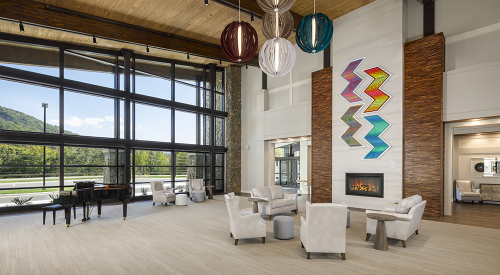 Community room at Tsali Care Center in Cherokee, North Carolina, featuring vaulted wood ceilings, a fireplace, colorful art, lounge seating, and large windows with mountain views.